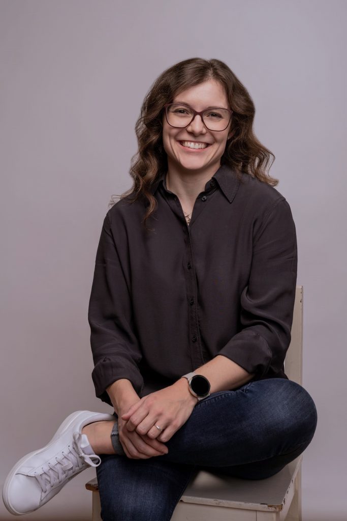 Eva Brucksch Ein freundliches Porträt einer Frau (Eva Brucksch) mit einer Brille und mittellangem, braunen, lockigem Haar, die auf einem Stuhl sitzt. Sie trägt eine dunkle Bluse, eine dunkelblaue Jeans und weiße Sneakers. Dazu trägt sie eine Smartwatch mit beigem Armband, eine silberne Kette und einen silbernen Ring. Sie hat ihr linkes Bein auf ihrem rechten abgelegt, die Hände liegen locker auf dem linken Unterschenkel. Der Stuhl ist aus weiß lackiertem Holz, der Lack splittert an einigen Stellen etwas ab. Der Hintergrund des Fotos ist hellgrau.