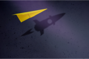 Leistung und Motivation Ein gelber Papierflieger vor einem dunkelblauen Hintergrund. Der Hintergrund ist mit dunkleren Sprenklern überzogen. Der Papierflieger wirft einen Schatten in Form einer Rakete.