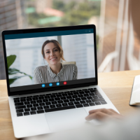 Ein Laptop (ein silbernes MacBook mit schwarzer Tastatur) zeigt eine Videokonferenz mit einer lächelnden Frau in gestreifter Bluse. Der Laptop steht auf einem Tisch in Buche-Optik. Rechts ist der Rand eines Notizbuchs erkennbar. Eine Person hat ihre rechte Hand neben das Mauspad des Laptops gelegt.