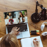 Online Training Eine Person mit dunkelblonden Haaren sitzt vor einem Laptop und nimmt an einem virtuellen Meeting mit vier Teilnehmenden teil. Unter dem Laptop liegt aufgeschlagen ein Notizbuch mit weißen, linierten Seiten. Neben dem Laptop liegt eine Brille mit runden Gläsern, darüber steht eine schwarze kleine Lampe im Retro-Stil. Darunter liegen silber-weiße Kopfhörer sowie ein neon-gelber und ein neon-orangener Textmarker. Von der Person vor dem Laptop sieht man die Oberseite des Kopfes, die linke Hand, die auf der Tastatur des Laptops liegt und die rechte Hand, die einen weißen Stift hält und auf dem Notizbuch liegt. Der Hintergrund ist ein Holztisch. Die Teilnehmenden des Meetings wirken positiv. Die Person links oben hält einen nach oben gestreckten Daumen in die Kamera. Die Person rechts oben lächelt in die Kamera und hat die linke Hand wie zum Winken erhoben. Die Person links unten strahlt in die Kamera und hält beide Hände ins Bild. Die Person rechts unten lächelt ebenfalls in die Kamera und deutet mit dem ausgestreckten Zeigefinger der rechten Hand ins Bild.